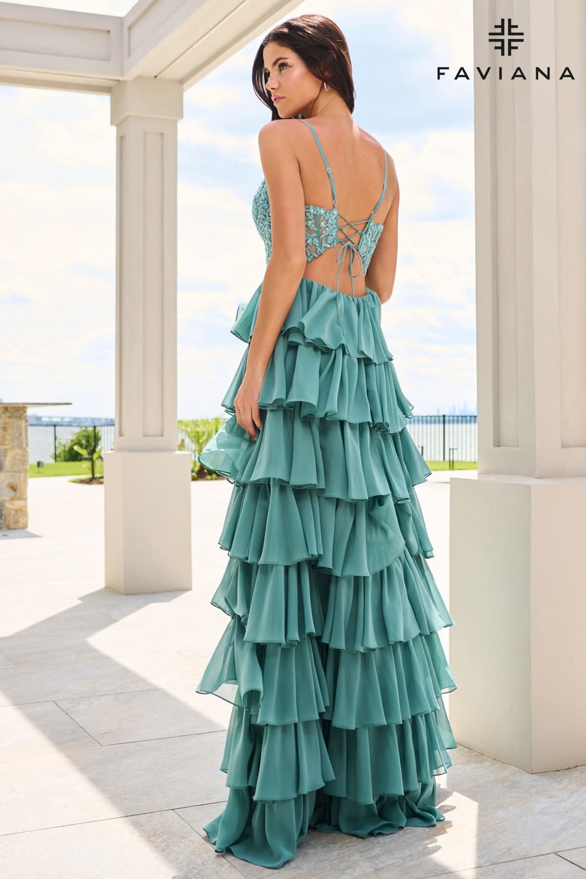 A woman wearing a teal green tiered gown with a lace-up open back, photographed from behind in an outdoor patio setting with columns. The FAVIANA brand logo is visible in the top right corner. A woman wearing a teal green tiered gown with a lace-up open back, photographed from behind in an outdoor patio setting with columns. The FAVIANA brand logo is visible in the top right corner.