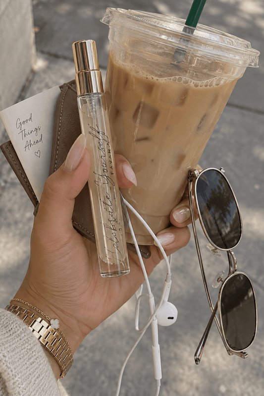 A hand holding a clear glass perfume bottle with a gold cap next to an iced coffee, sunglasses, white earbuds, and a brown wallet on a sidewalk.