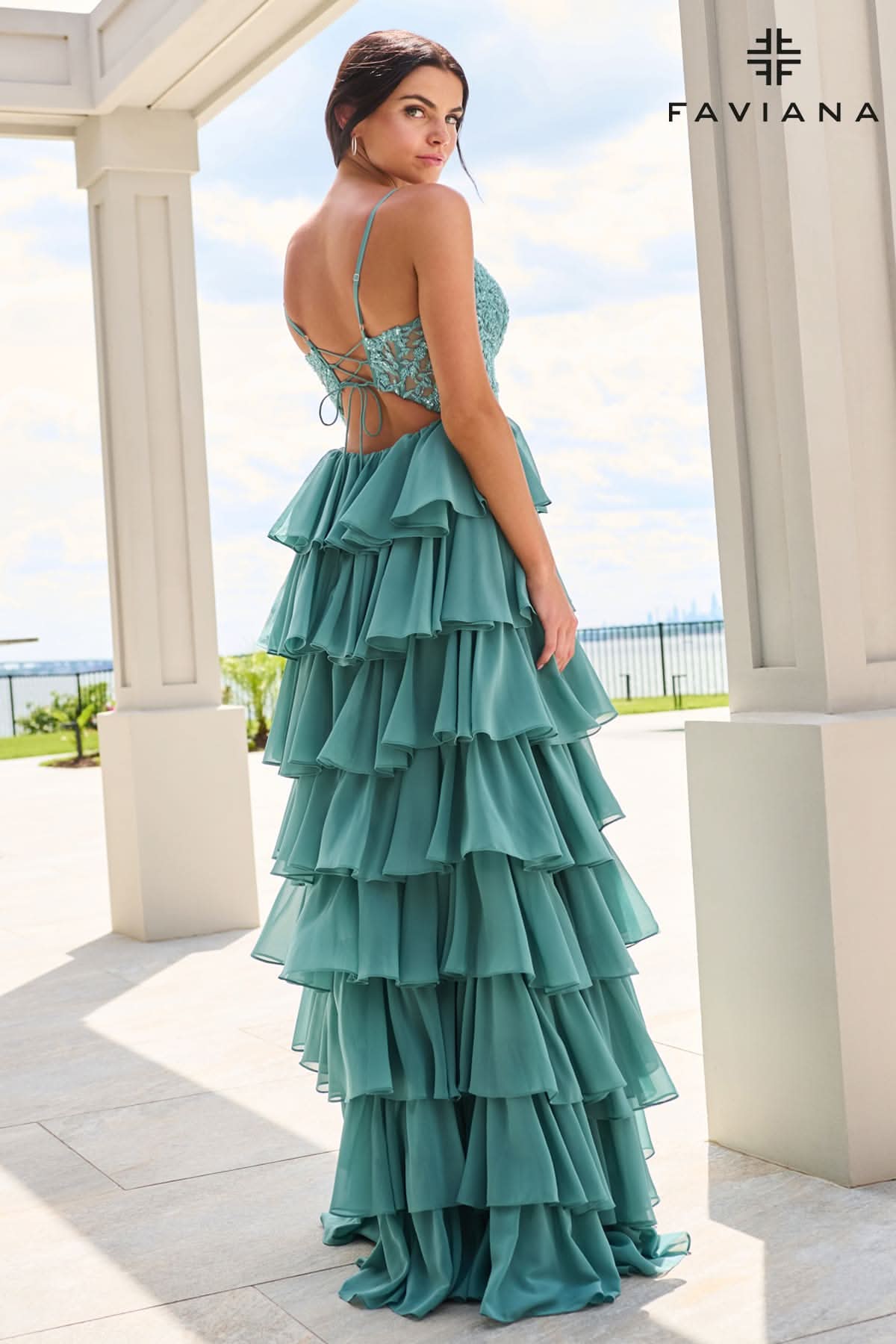 A woman modeling a teal green tiered ruffle maxi dress with a lace-up open back, standing on a tiled patio under white columns.