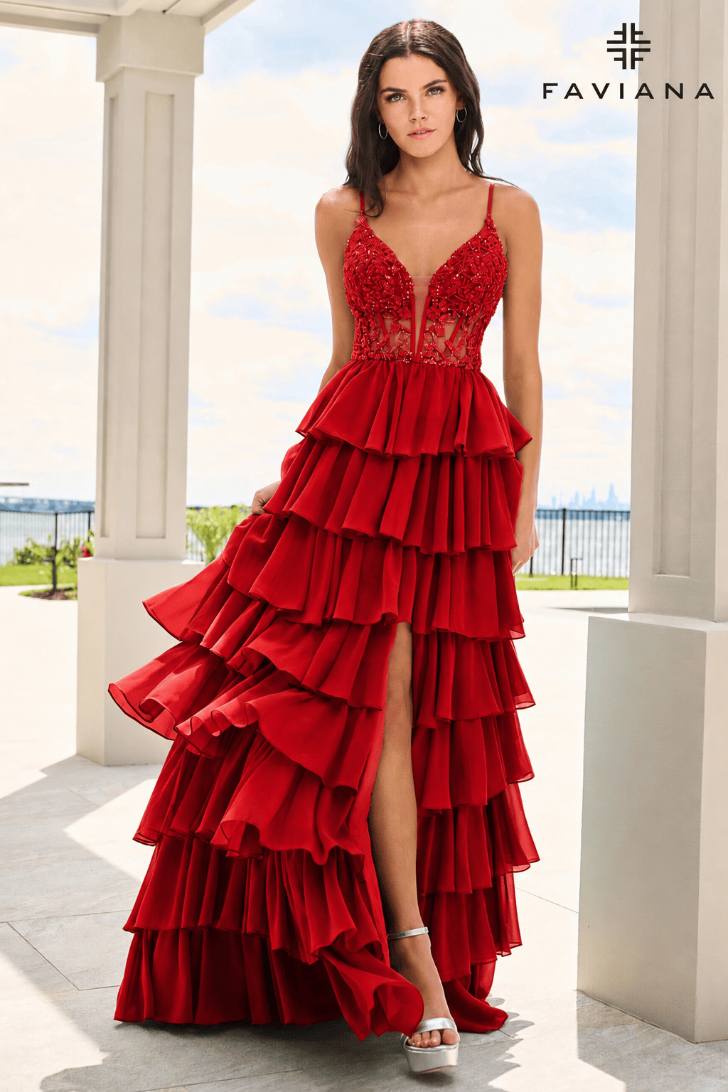 A woman models a bright red formal gown with a sheer lace bodice, thin straps, and a dramatic multi-tiered ruffled skirt with a thigh-high slit, standing in a sunlit outdoor porch setting. The FAVIANA brand logo is visible in the top right corner.